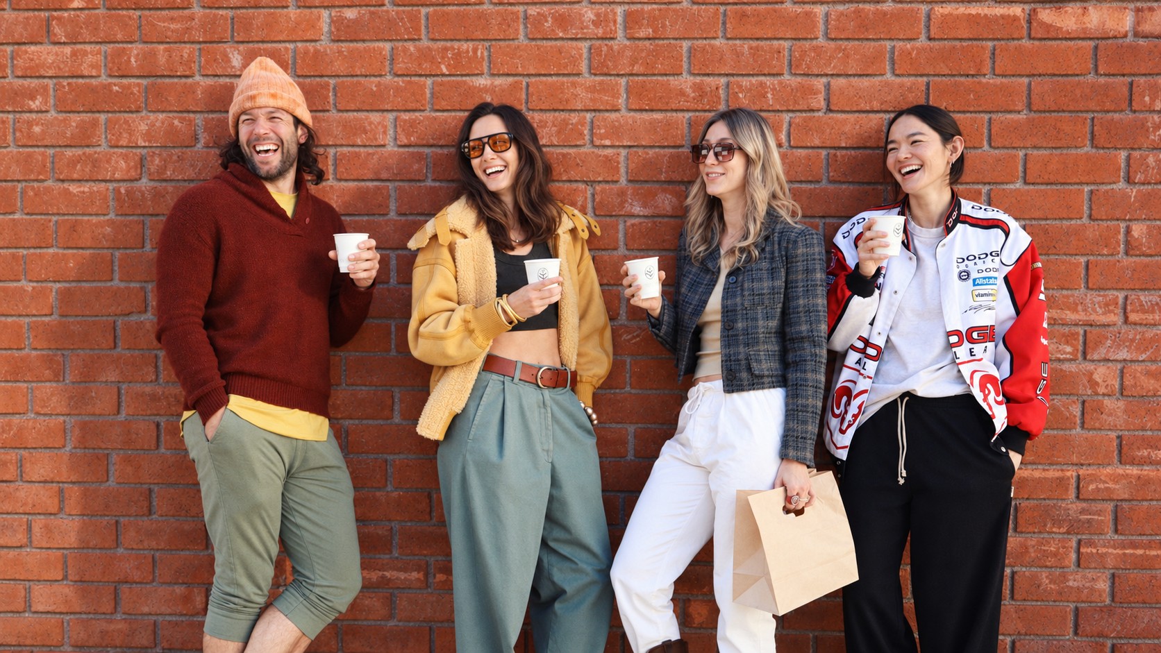 Four friends laughing against a brick wall, coffees from Cherish in hand
